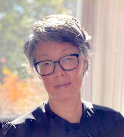 A woman in a dark shirt, with short hair and glasses stands i front of a window overlooking a garden.