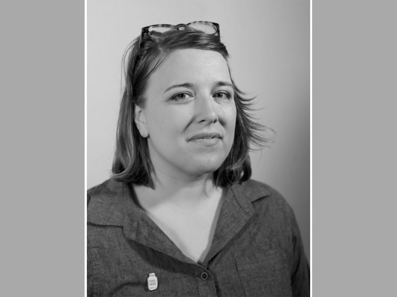 Black and white headshot of Kelsey Halliday Johnson, a white woman with shoulder length hair, eyeglasses pushing her hair back, wearing a dark gray button-up shirt.