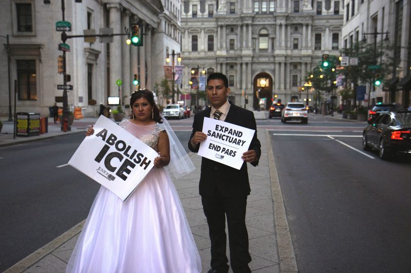 A man and women with brown skin, dressed in a suit and bridal gown, stand in the center of Broad street in front of Philadelphia City Hall, holding signs that say "ABOLISH ICE" and "EXPAND SANCTUARY / END PARS"