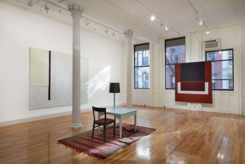 Installation view of a white-walled, wooden-floored gallery, with two large abstract paintings on the two walls, and a desk with a chair in the center of the room.