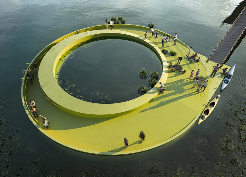 A green circular platform with a circular hole in the middle sits atop a body and is connected to land by a black ramp. of water. People stand on the platform.