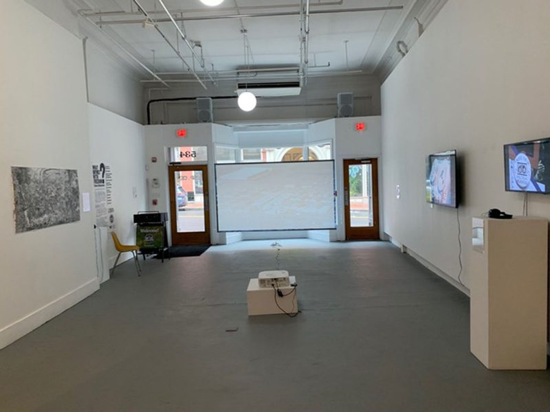 Gallery in SPACE, with a video projected onto a screen in front of bay windows between two entrances to the space, and various hanging drawings and video artworks on monitors.