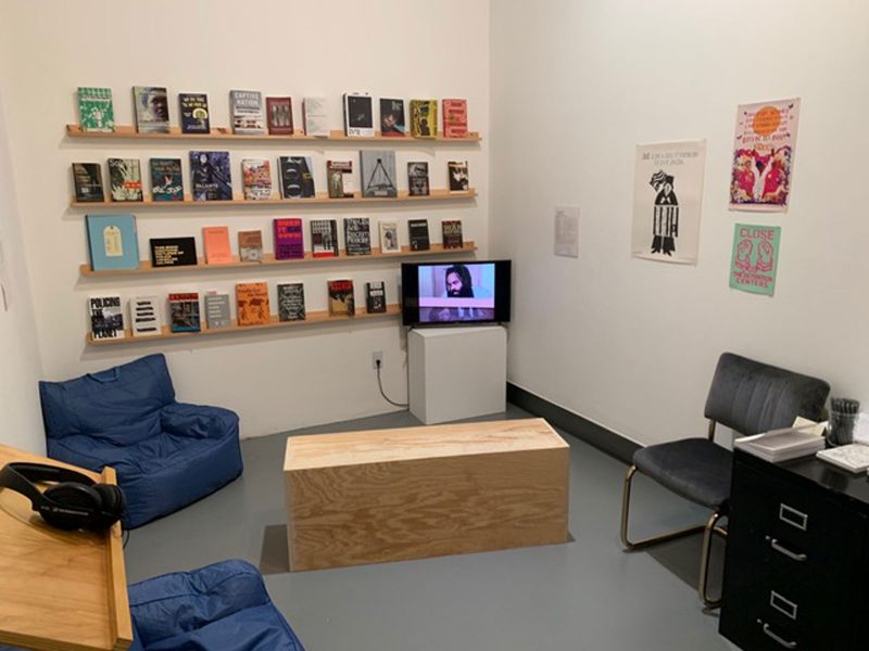 Sitting space with two beanbag chairs and another metal and fabric chair, surrounding a low wooden table, with a video playing on a nearby podium, and floating shelves displaying various books. 