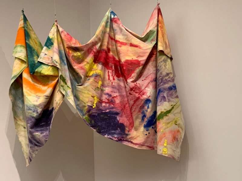 Colorful abstractly painted fabric draped in midair in the corner of a gallery space, suspended at four points with fishing wire.