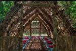 Red pillows on blue mats line the interior edges of a light-filled outdoor structure made out of organic material (indigenous weed, phragmites), in a lush green environment