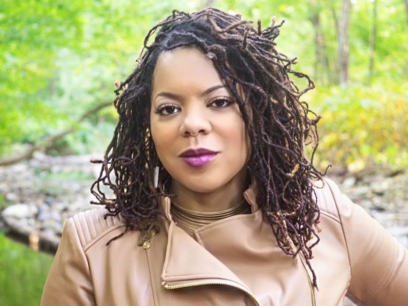 A Black woman with dark shoulder-length micro braids, wearing purple lipstick and a tan leather jacket, smiling.