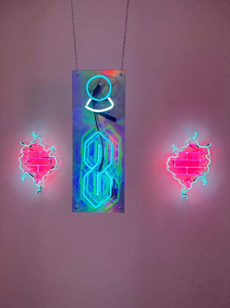 Neon sculpture of the "Cool S" (a popular stylized S) and a circle on a piece of metal; on either side are identical but mirrored neon sculptures of blue squiggly lines inside of which are red brick patterns. 