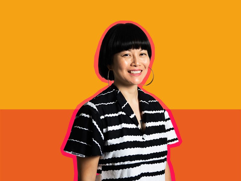 A woman with black hair, big hoop earring a black and white blouse, smiles warmly at you. She is outlined in orange and the background is two shades of orange, one light and the other darker.