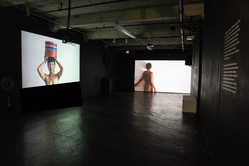 Photograph of a dark space with a black-painted floor shows two large videos projected onto two walls, both featuring a woman. 