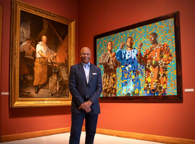 A Black man with a shaved head wearing a dark blue suit stands in front of two paintings, one old (left; a portrait of a white blacksmith) and one new (right; portrait of three black men with decorative patterned background)