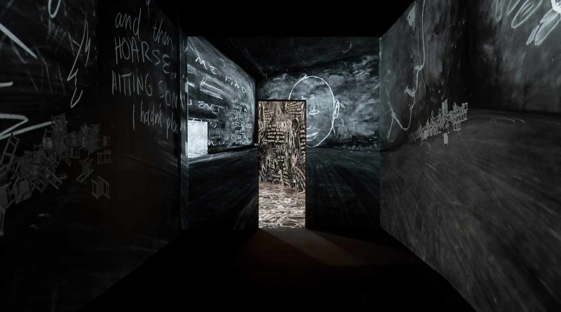 Installation view of a dark corridor with black floors and dusty chalk black walls, with white chalk drawings of faces and words, smudged in many areas, leading to another similar room with many more white drawings on both the walls and floor.