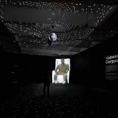 Larger than life sized projection of a Black man in an arm chair in the farthest corner, in a black room with a disc ball hanging in the center, casting white light on the ceiling and floor.