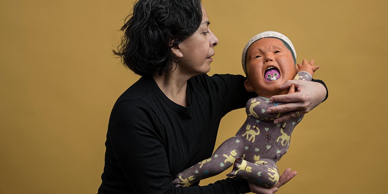 A photo shows a woman with black hair and wearing a black longsleeve t-shirt holding a crying baby with a toy caught in its mouth who seems to be wiggling to get out of her arms, and the background is a flat ochre color.