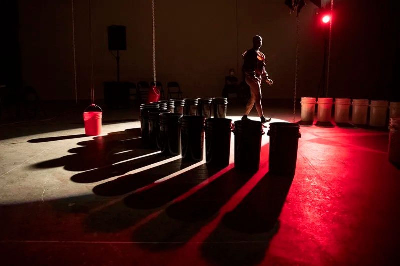 A red-lit stage with two sets of buckets-- a black set and a white set-- arranged in two semi-circles, in between which a figure is walking towards the center.