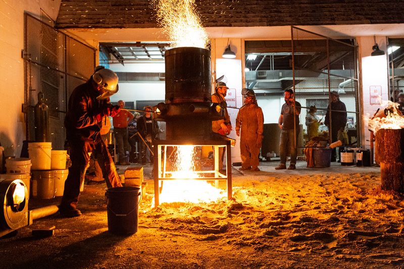 A man in head to toe protective gear operates a iron forge with sparks flying out of it, while people observe from a distance.