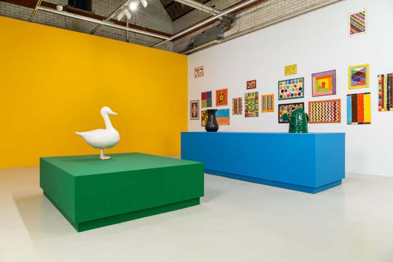 Gallery room with one white wall, with hanging abstract art on it, and one yellow wall, with two pedestals in the center of the room, one green with a white sculpture of a duck on it, one blue with a black pitcher and a green watering can on it.