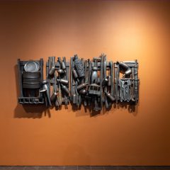 Metal sculpture hanging on an orange wall, shaped like a wooden fence, upon which are collaged metal objects like empty bottles, laundry pins, and wrinkled clothing.
