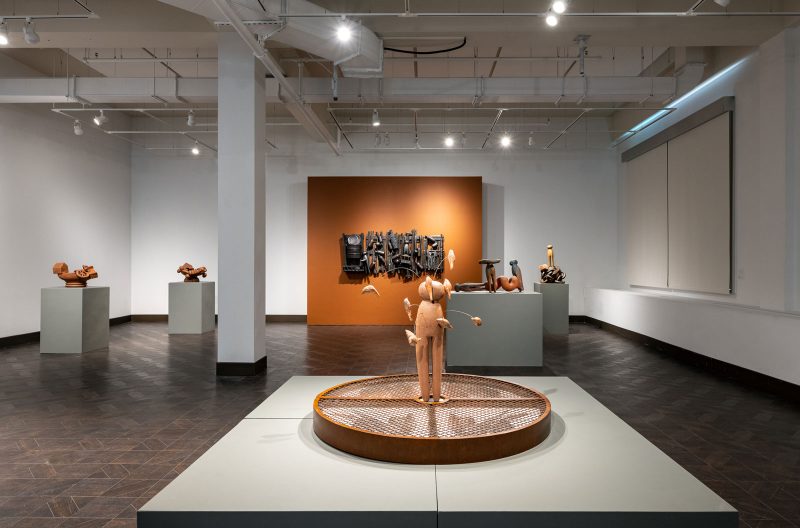 Installation view of a gallery room, with a large sculpture on a white pedestal in the foreground, a large wooden laundry pin with smaller wooden birds extending from it via flexible wire, in the center of a circular  metal grate, which is the base of the sculpture.