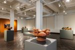 Installation view of a gallery room, with multiple gray pedestals upton which are stylized unglazed terra-cotta bowls, each with unique aspects that are related to the rural farms for which the bowls are a tribute.