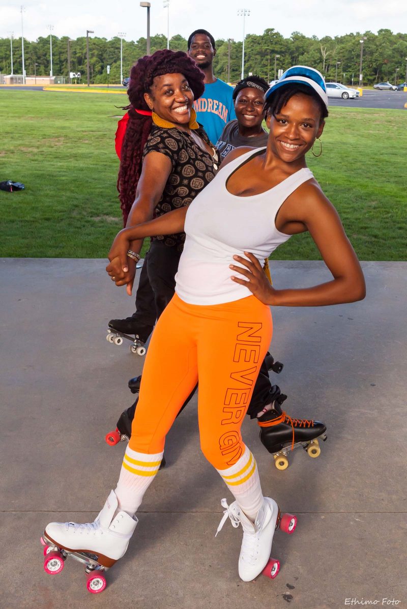Sanchel Brown, a Black woman with short braided hair wearing white and orange athletic clothing and roller skates, smiling and posing holding hands with four other Black people in roller skates, all standing in a line and smiling together.