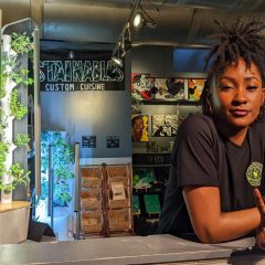 Christina, a Black women with medium-length black hair, leaning on a counter and holding up a peace sign in front of a hydroponic garden and art, primarily portraits, hanging on the wall behind them.