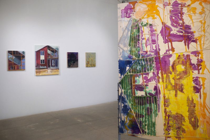 On the left, four paintings hang on a gallery wall, featuring scenes of barns, wood work, and trees at night and during the day; on the right an abstract colorful two-sided painting hangs on a metal stretcher, with purples, yellows, green, on raw canvas.