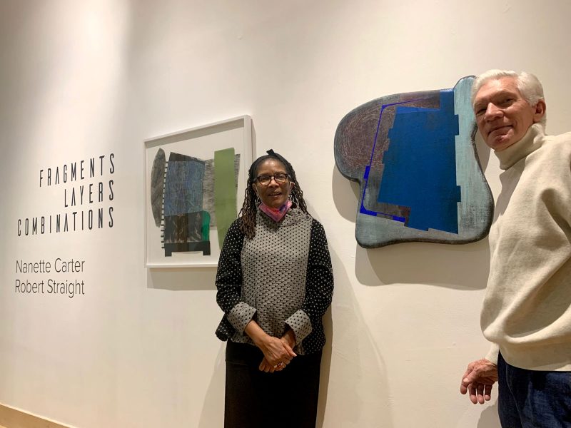 A Black woman (left) with collar bone-length braided hair, wearing a black and gray patterned shirt, and a white man (right) with ear-length white hair, wearing a tan turtleneck sweater, stand posing with their artwork in a gallery.