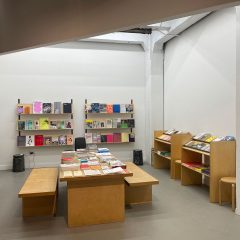 Book shop with simple wooden furniture: benches, tables, book shelves; with books and papers arranged on display on the table and shelves