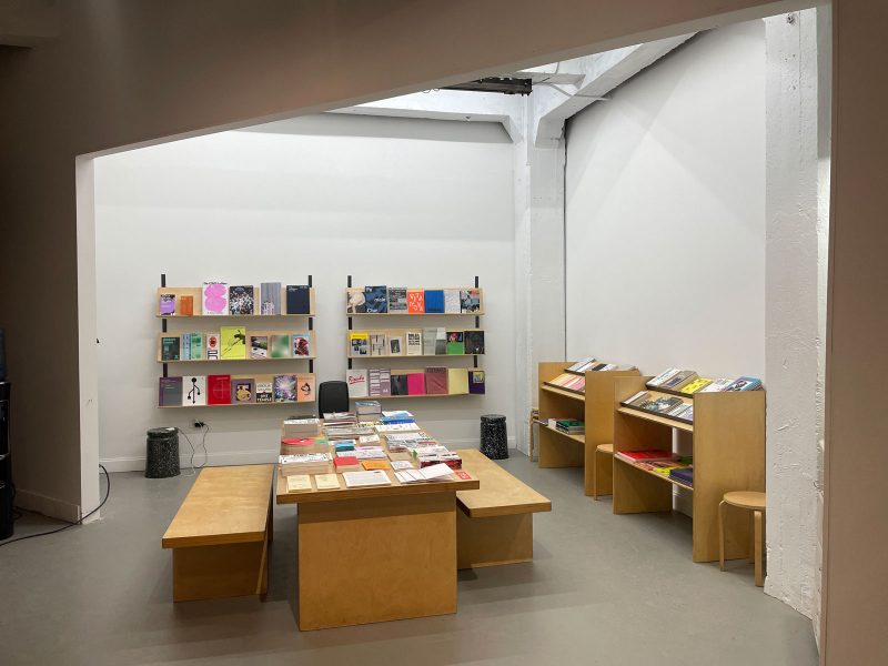 Book shop with simple wooden furniture: benches, tables, book shelves; with books and papers arranged on display on the table and shelves