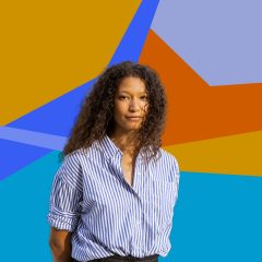 A woman with brown skin and long curly hair parted down the middle, wearing a blue and white striped blouse, with her hands behind her back, in front of a colorful decorative background.