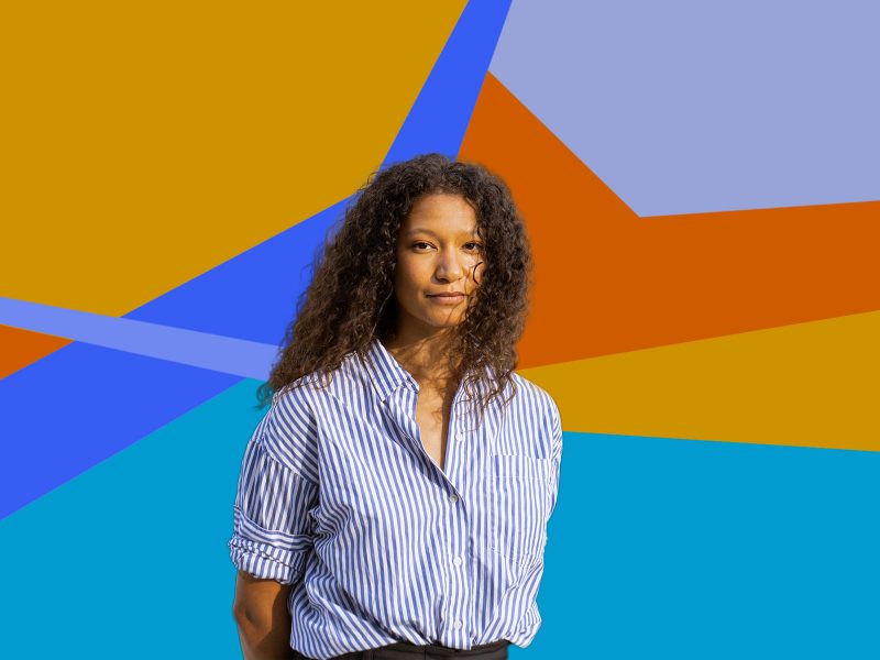 A woman with brown skin and long curly hair parted down the middle, wearing a blue and white striped blouse, with her hands behind her back, in front of a colorful decorative background.