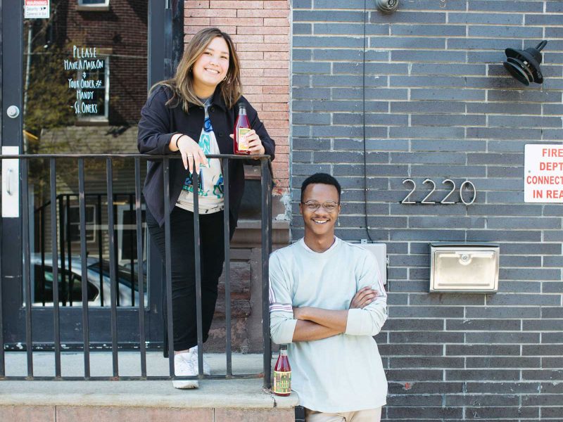 Alyssa Foronda, a Filipino woman with long brown and blonde hair, poses smiling & holding a bottle of JamBrü Kombucha; Jamaar Julal, a Black man with short black buzzed hair, stands leaning nearby with his hands across his chest, smiling next to a bottle of JamBrü Kombucha.