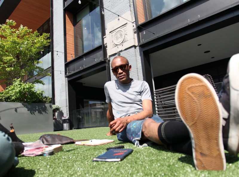 Claes Gabriel, a Black male artist wearing jean shorts, sunglasses, sneakers, and a gray t-shirt, sitting in the grass surrounded by his journals/ sketch books, and looking into the camera.