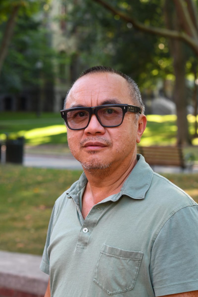 Color photo of a man, Ken Lum, outside in a park, surrounded by trees and grass and wearing a pale green shirt and tinted-glass, black glasses, looking up pensively.