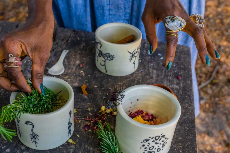 Close-up on the hands of a Black woman as she takes a sprig of what looks like pine from a ceramic white cup with black illustrations on it that's sitting on a wooden table.