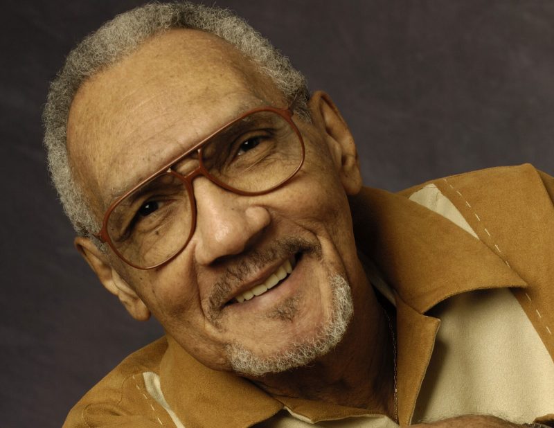 Bob Perkins, a Black man with short salt and pepper hair and facial hair, wearing brown aviator-shaped glasses, a tan and cream colored collared shirt, smiling in front of a gray mottled photo background.