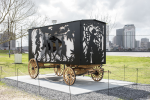 Carriage whose four walls are cut out in intricate narrative artwork featuring silhouettes of different people and actions on each wall, installed outside of the National Gallery of Art on a marbel slab.