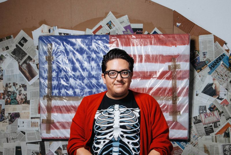 Justin Favela, a Latinx artist with short black hair combed back and to the left, wearing a black shirt with a skeleton on it, black glasses, and a red cardigan, smiling in front of a painting of the American flag wrapped up in plastic, hanging in front of newspapers pasted in every which way on the wall on cardboard.