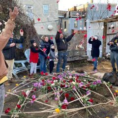 A group of people laughing and throwing flower petals in the air above a small pit in the ground that has many sticks laid across it that are decorated with flowers.