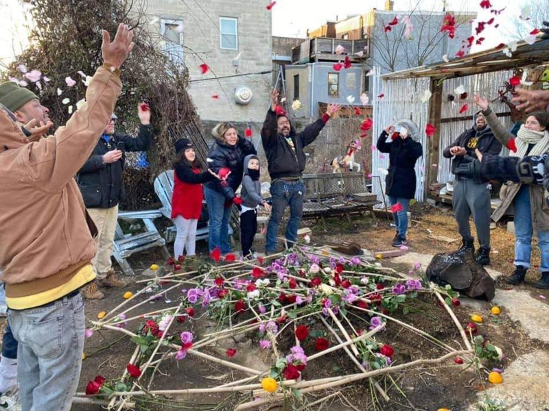 A group of people laughing and throwing flower petals in the air above a small pit in the ground that has many sticks laid across it that are decorated with flowers.