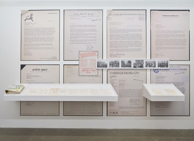 Gallery exhibition featuring many blown-up and framed documents from the Comprehensive Employment and Training Act (CETA), detailing various organizations who participated. Also attached to the wall are glass cases with more, standard-sized documents, and a series of 6, small, black & white photos that are CETA-related.