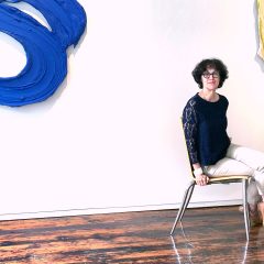 Christine Pfister, a white woman with wavy hair cut at a chin-length bob with short bangs, wearing a navy blue lace blouse, tan chino pants, and light brown flats, sitting on a wooden chair with metal legs in Pentimenti Gallery. The gallery has dark and light brown hardwood floors, white walls, and partially visible on the walls are hanging sculptures, depicting abstracted and enlarged paint brush strokes in vibrant colors.