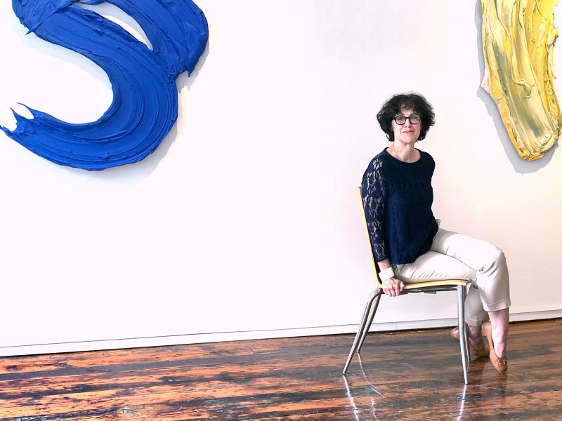 Christine Pfister, a white woman with wavy hair cut at a chin-length bob with short bangs, wearing a navy blue lace blouse, tan chino pants, and light brown flats, sitting on a wooden chair with metal legs in Pentimenti Gallery. The gallery has dark and light brown hardwood floors, white walls, and partially visible on the walls are hanging sculptures, depicting abstracted and enlarged paint brush strokes in vibrant colors.