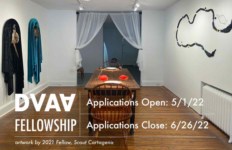 White text announcing DVAA's open call for Fellowship applications through 6/26/22, overlaid over an art installation of a wooden table with bodily sculptures on them in the center of the table, plus garments and wall sculptures hanging on the adjacent walls.