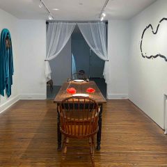 An art installation of a wooden table with bodily sculptures on them in the center of the table, plus garments and wall sculptures hanging on the adjacent walls.