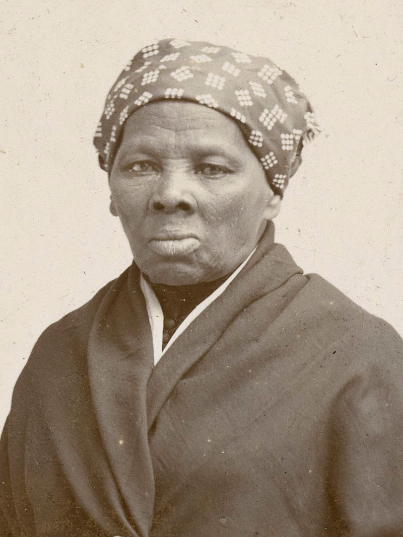 Photograph portrait of Harriet Tubman, a Black woman wearing a brown or gray robe over a black blouse, and a dark scarf with white patterns wrapped around her hair.