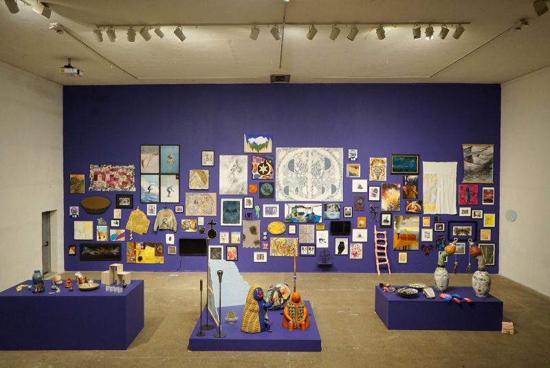 Installation view of a gallery room with one wall painted indigo, on which there are nearly 100 2D artworks hung; and three indigo platforms, on which there are a number of sculptures, approx. 20.