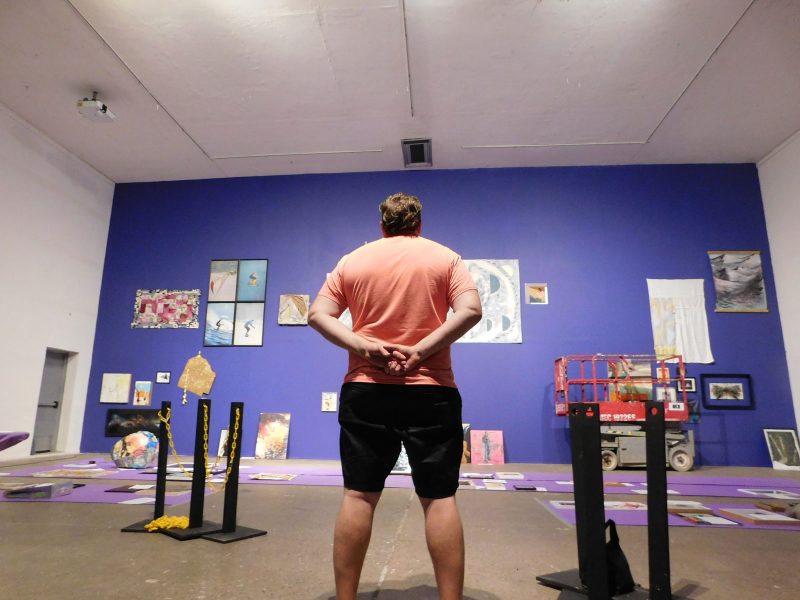 A white man with curly blonde-brown hair, wearing a coral-colored shirt and dark shorts, pictured from behind, holding his hands behind his back and looking at a purple gallery wall with artwork hung on it, and other artwork to be hung sitting on purple paper on the floor below.