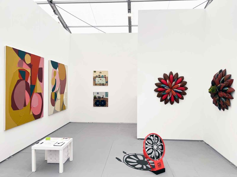 Installation view of artwork in a gallery space with white walls and gray floors: on the left wall; two large abstract paintings of colorful geometric shapes; on the farthest back wall, two small works of perspective-warped interior spaces; on the two L-shaped walls, two hanging 3D sculptural fiber works of flowers; and on the floor, a sculpture of a hubcap with a rendered black silhouette depicting a shadow on the floor behind it.