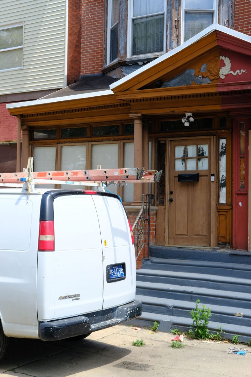 Rowhouse with blue steps leading up to a wooden door; four windows are to the left of the door; the awning of the home is painted half orange and half red, and there is a bay window that juts out on the second floor, and the windows are boarded up. A white work truck with a ladder strapped to the top is parked on the curb next to the blue steps.